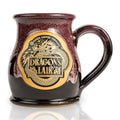 The front of the Bones Coffee Company Dragon’s Lair hand thrown mug with the Dragon’s Lair art on the golden medallion. The mug is black colored with a burgundy and white glaze on top.
